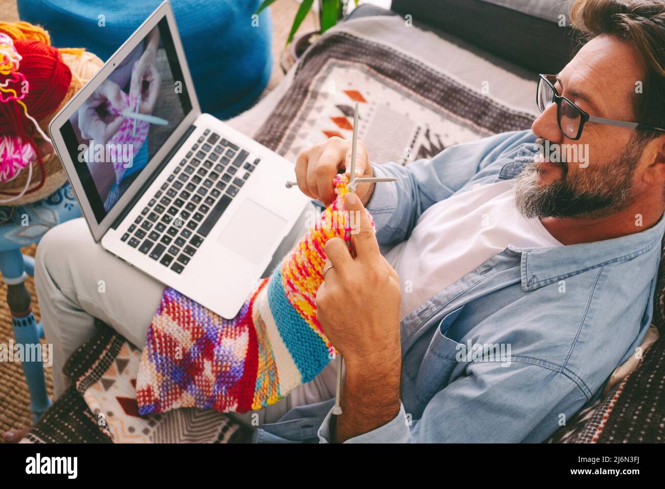 Handsome adult man learning how to knitting work with woolen yarn at home watching an online class tutorial on laptop computer. Concept of female Stock Photo