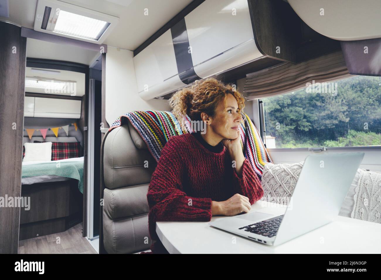 Adult woman use laptop computer inside a camper van recreational vehicle sitting at the table with bedroom in background and nature park outside the Stock Photo