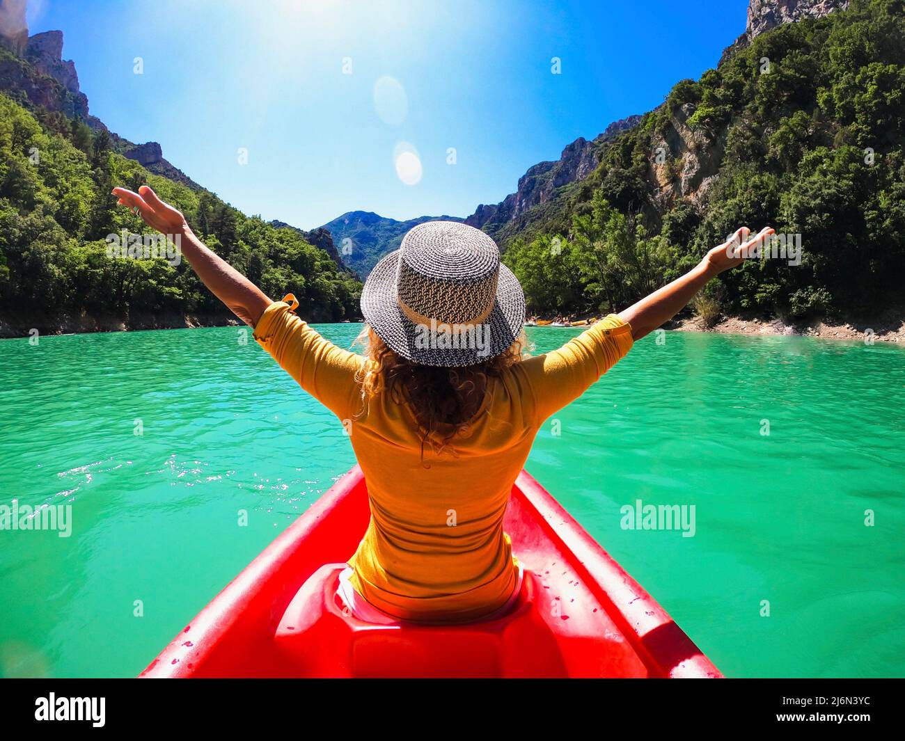 Rear view of woman in hat sitting on kayak with her arms raised on a beautiful lake between mountain. Woman enjoying kayaking sailing on lake water Stock Photo