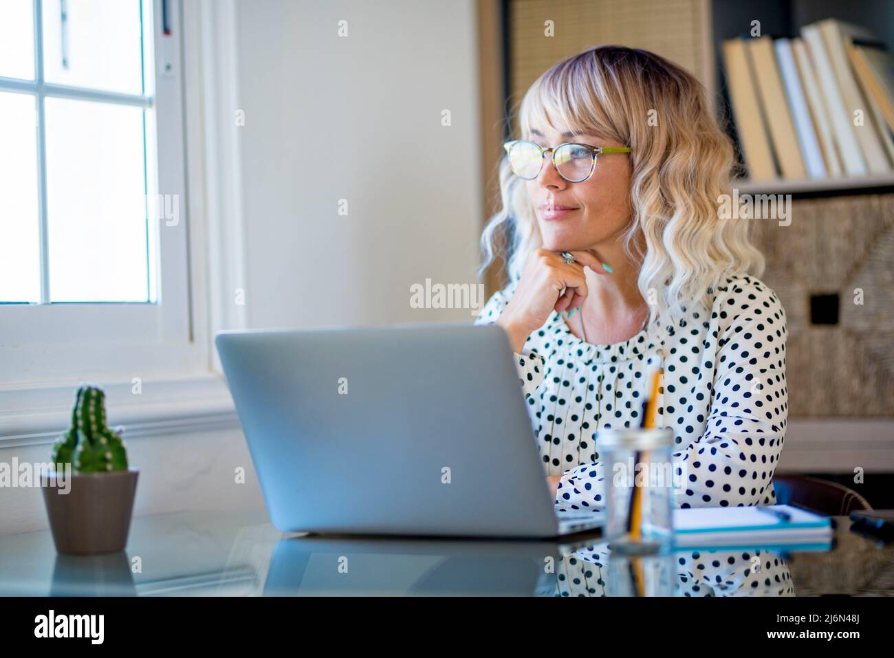 Modern web worker adult woman rest and have break relax time at the desktop in home office workstation looking outside the window and thinking. Happy Stock Photo
