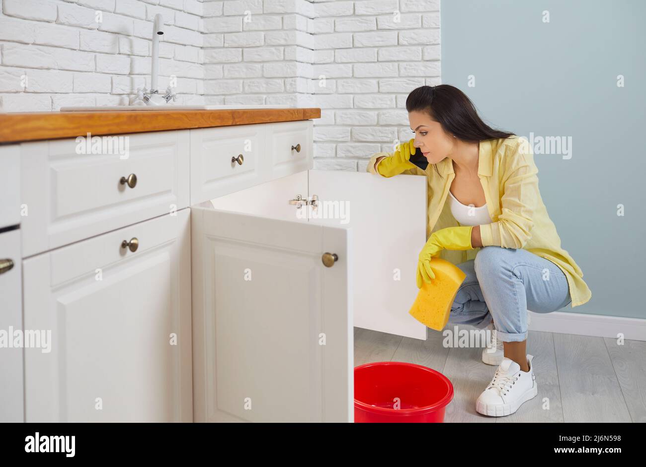 Frustrated woman on phone calls plumber because of leaking pipe in sink in kitchen. Stock Photo