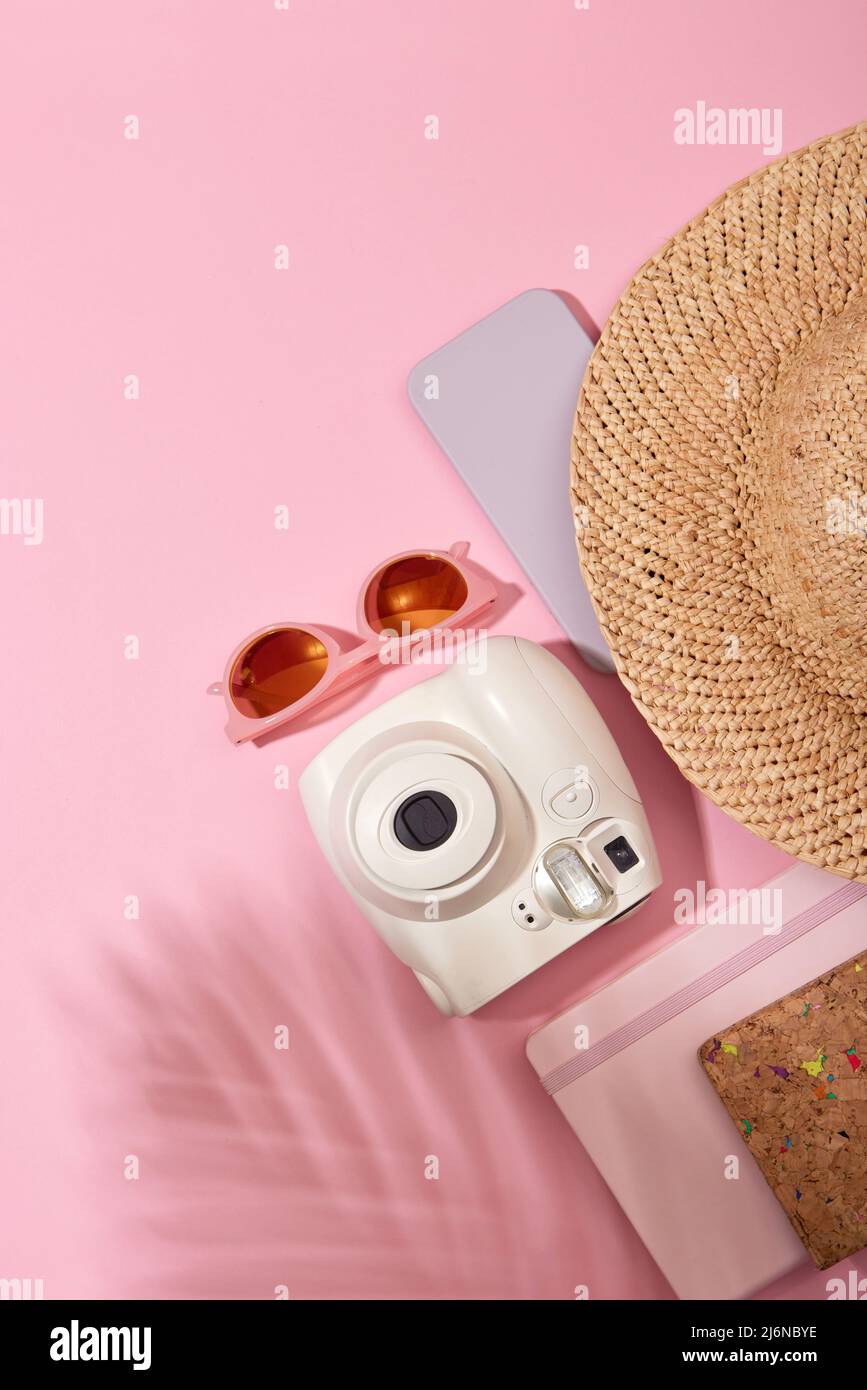 From above of instant photo camera with sunglasses and straw hat placed near notebook on pink background with shadow of plant Stock Photo