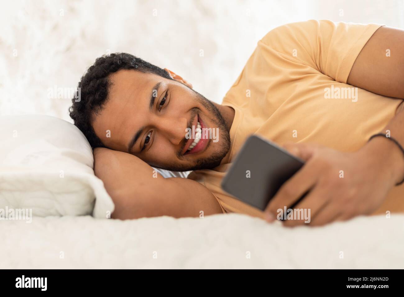 Happy Arabic Guy Texting Via Phone Lying In Bedroom Stock Photo