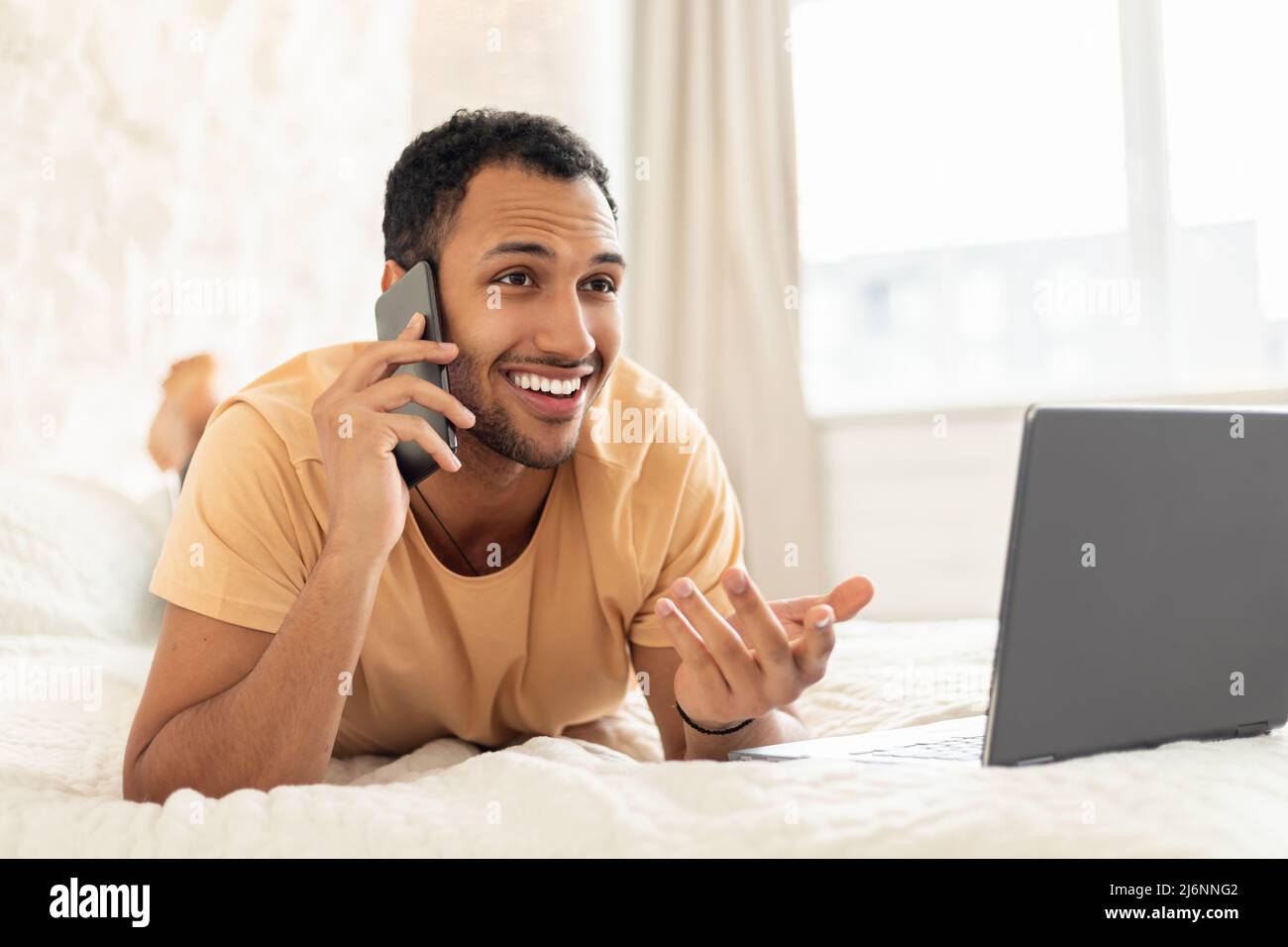 Middle Eastern Guy Talking On Cellphone Using Laptop In Bedroom Stock Photo