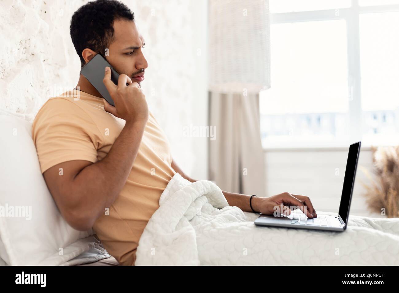 Arabic Guy Talking On Phone Using Laptop Working In Bedroom Stock Photo