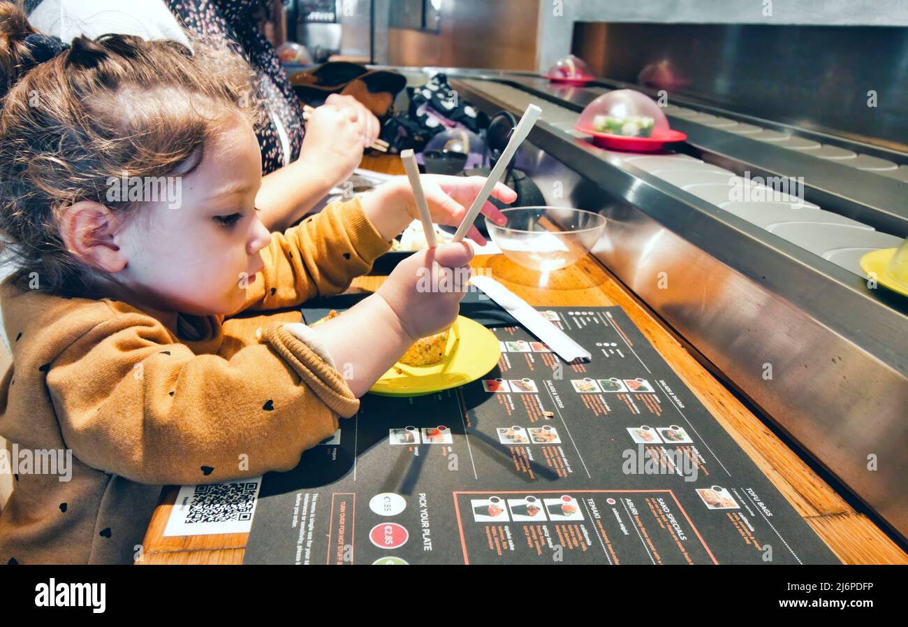 Cute little girl using chopsticks to eat sushi at a restaurant with self-service conveyor belt Stock Photo
