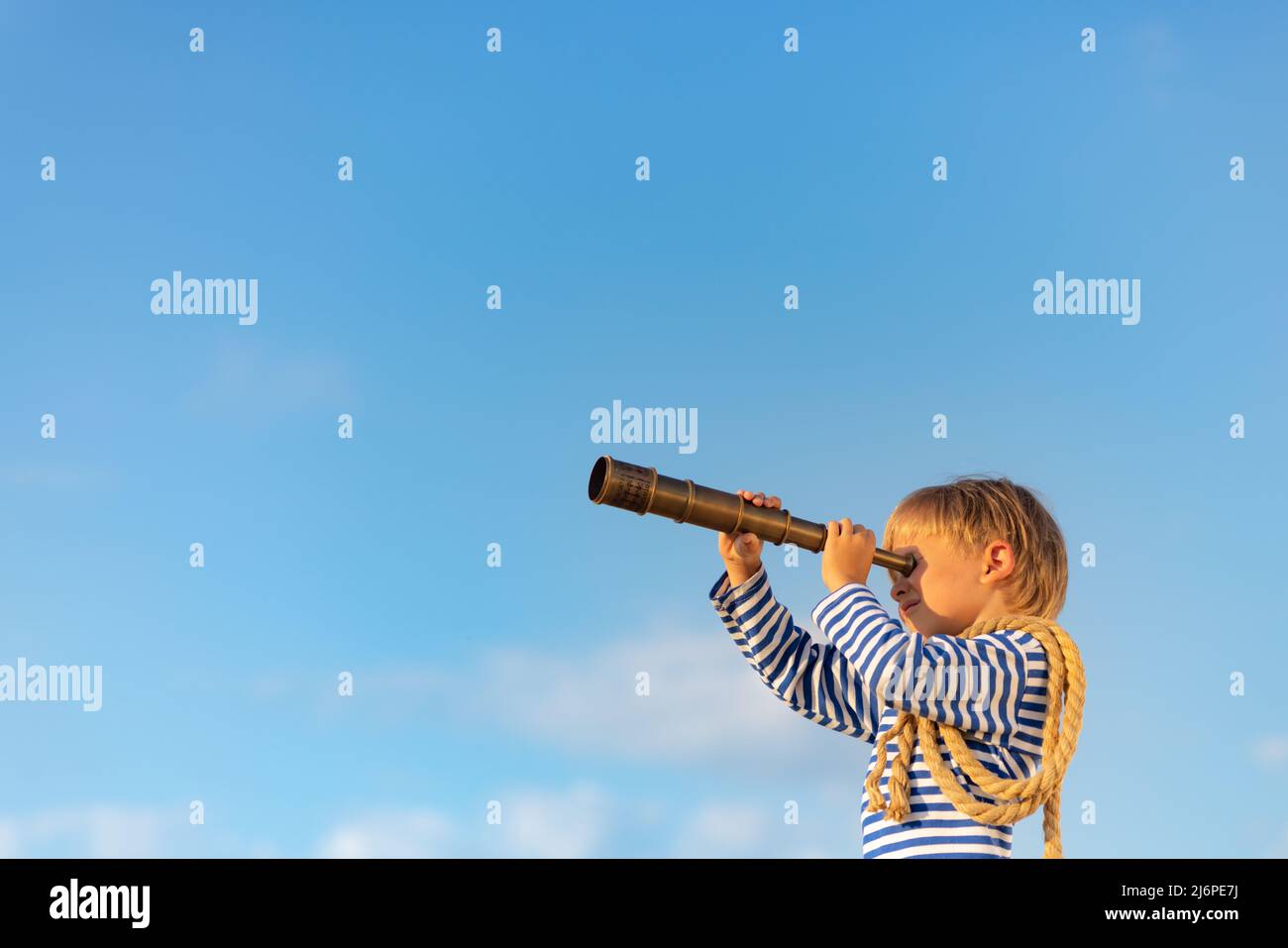 Happy child looking through vintage spyglass against blue sky. Kid having fun in summer. Imagination and freedom concept. Stock Photo