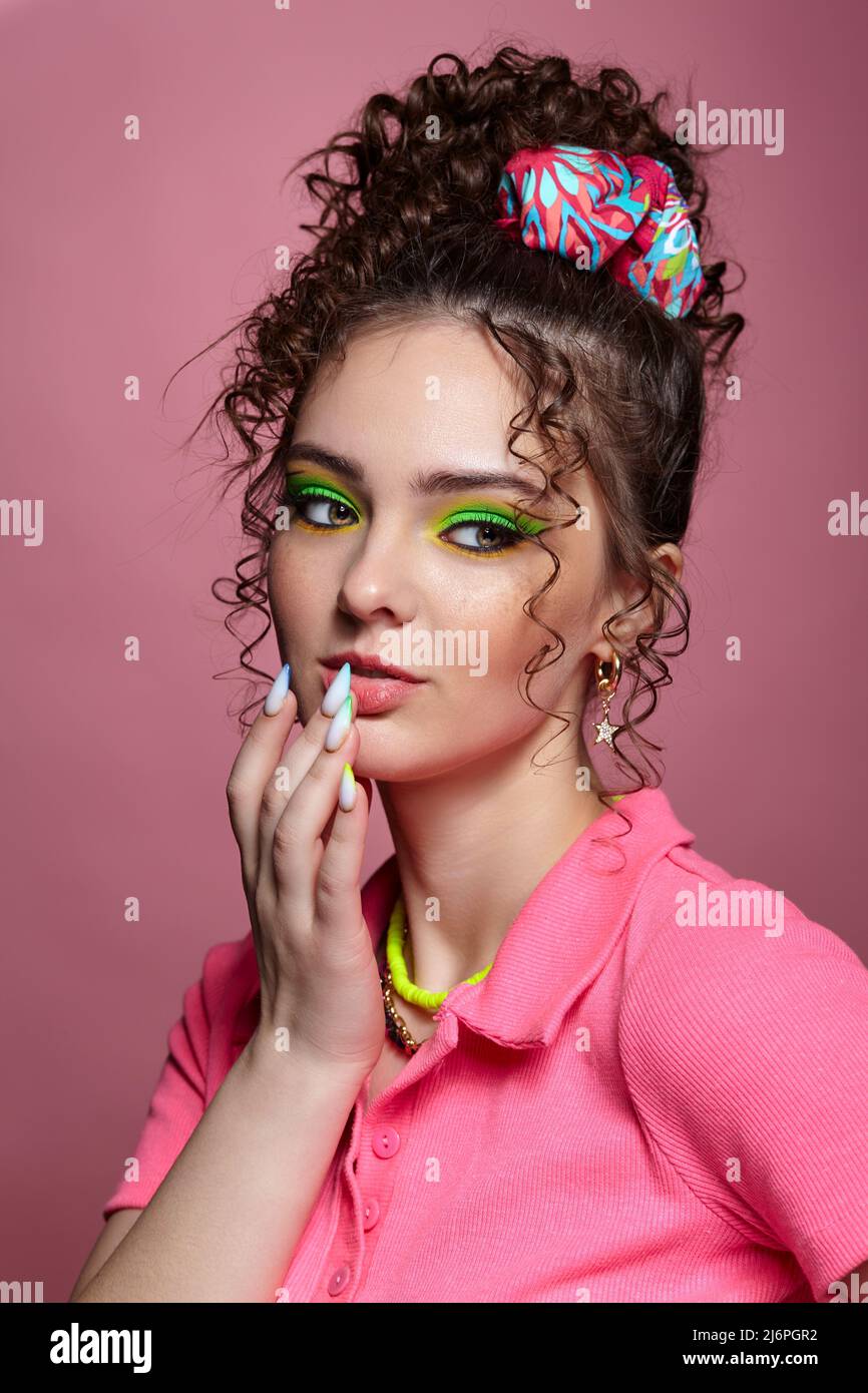 Portrait of young woman on pink background. Female with unusual green eyes shadows makeup, curly hair and earrings. Hand is near lips. Stock Photo