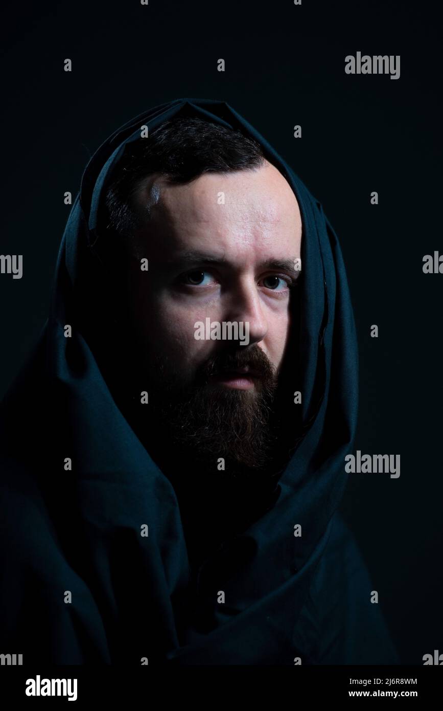 dramatic religious portrait of a bearded guy in a black cape on a dark background. Stock Photo