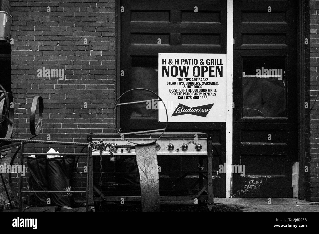A sign reads H & H Patio & Grill Now Open hanging above an outdoor grill and trash outside of a closed restaurant. Captured on analog black and white Stock Photo