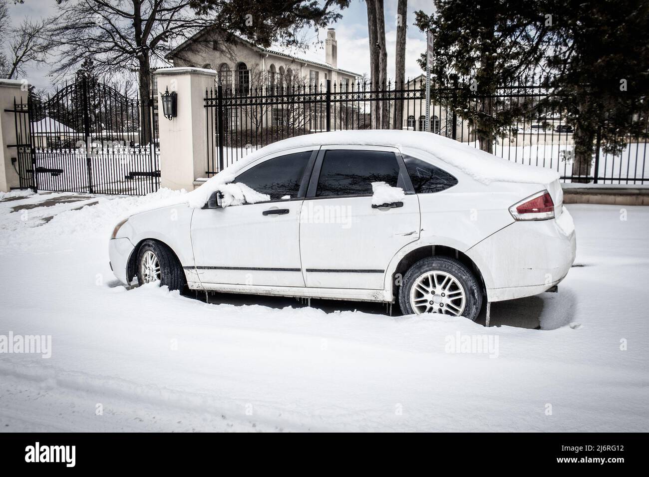 Dirty white car with scuffs parked in deep  snow on residential street outside tall fence with luxury mansion and estate in background. Stock Photo