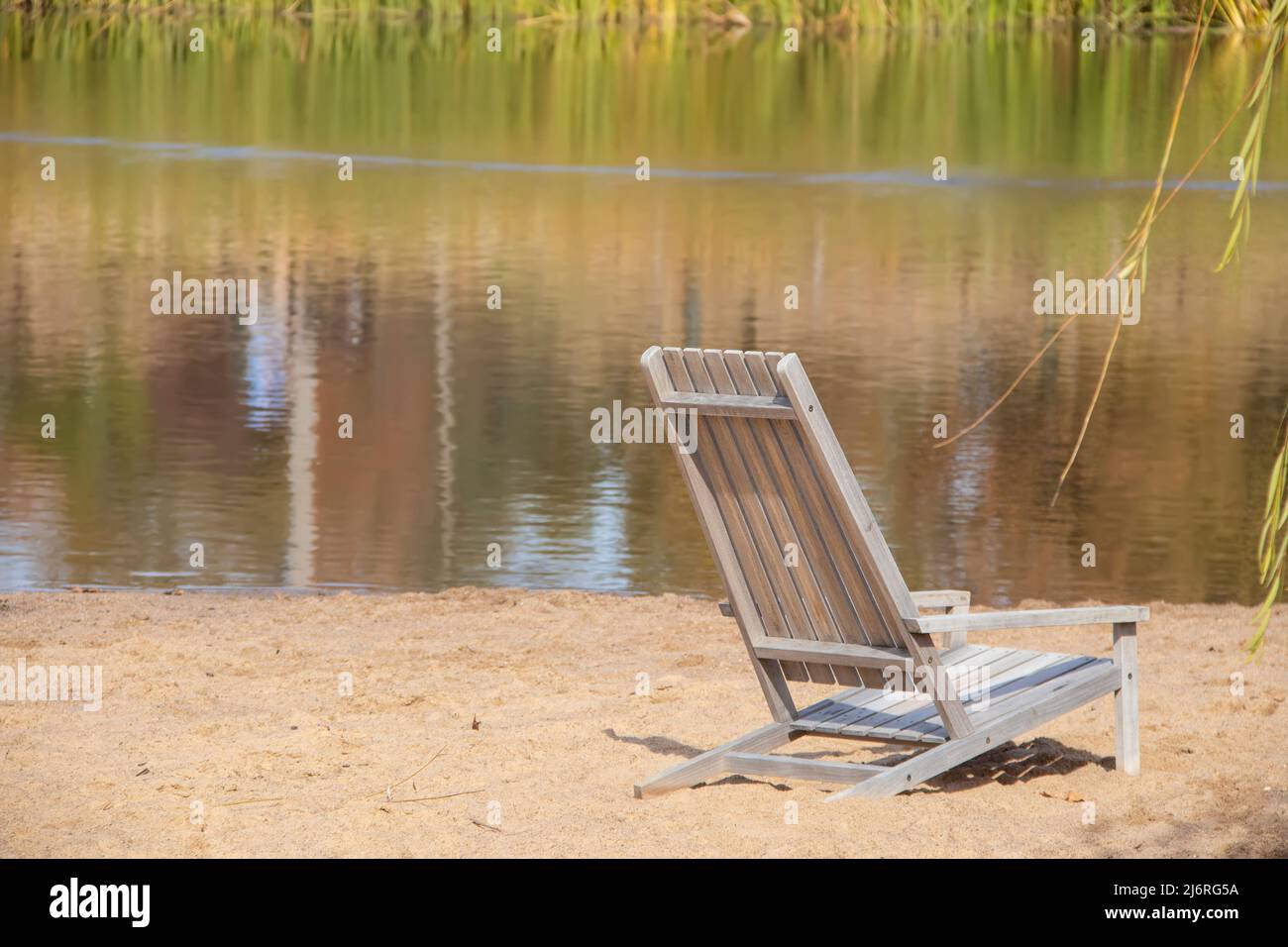 Dreamy scene of wooden beach chair in sand with reflections of reeds in water and willow branches on side - selective focus. Stock Photo