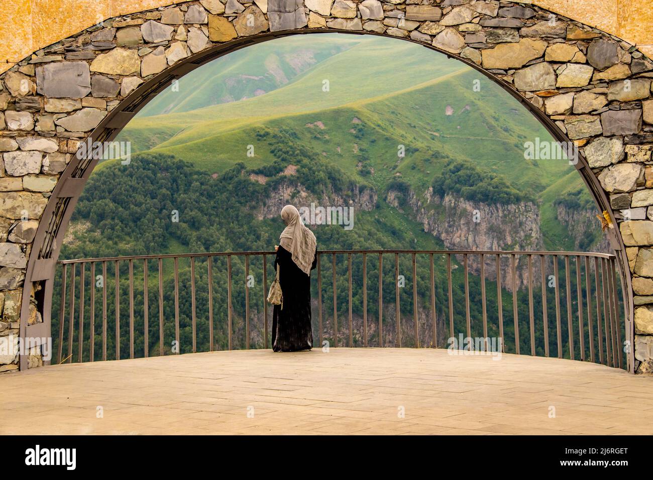 Female tourist in Abaya and Hijāb checks phone standing in arch of Georgia Friendship Monument on Military Highway with beautiful mountain view behind Stock Photo