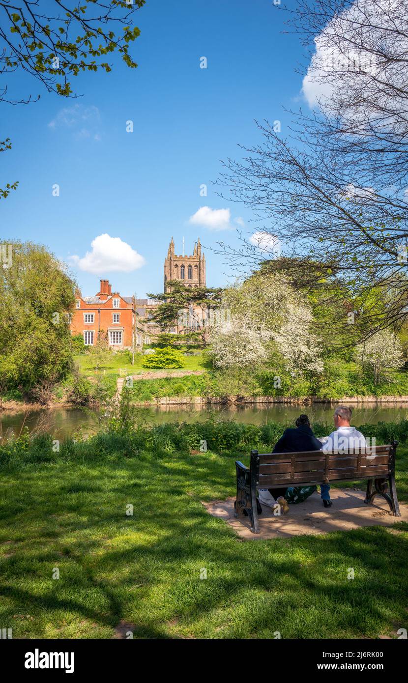Two adults sitting on a park bench over looking Hereford Cathedral from the banks of the River Wye, Hereford, England, UK Stock Photo