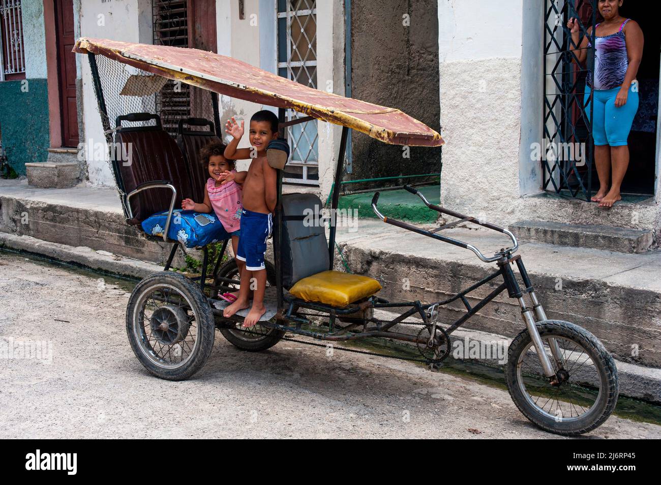 Young kids wave at the camera standing on a bicycle in front of their mother in Havana, Cuba. Stock Photo