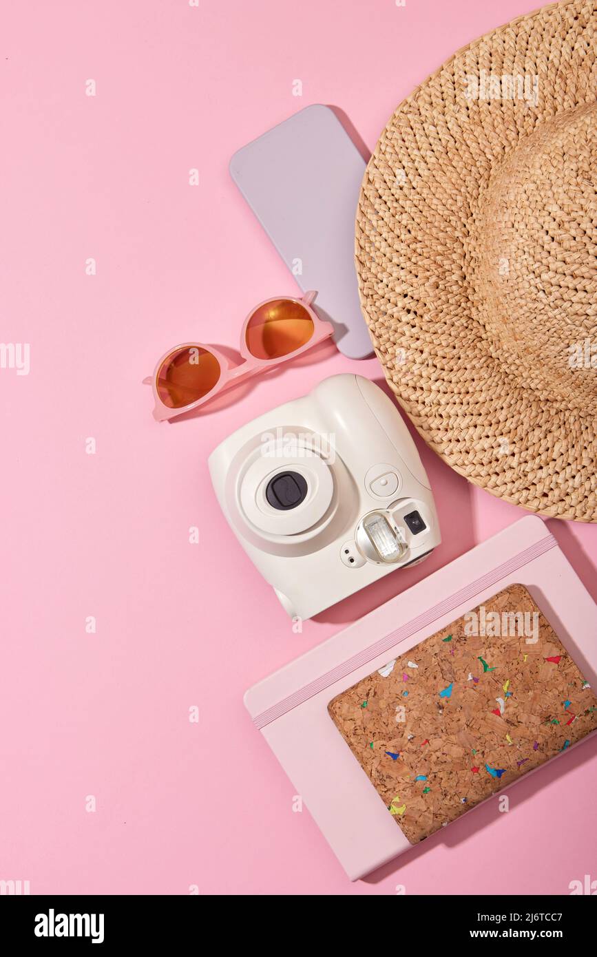 From above of instant photo camera with sunglasses smartphone and straw hat placed near notebook on pink background Stock Photo