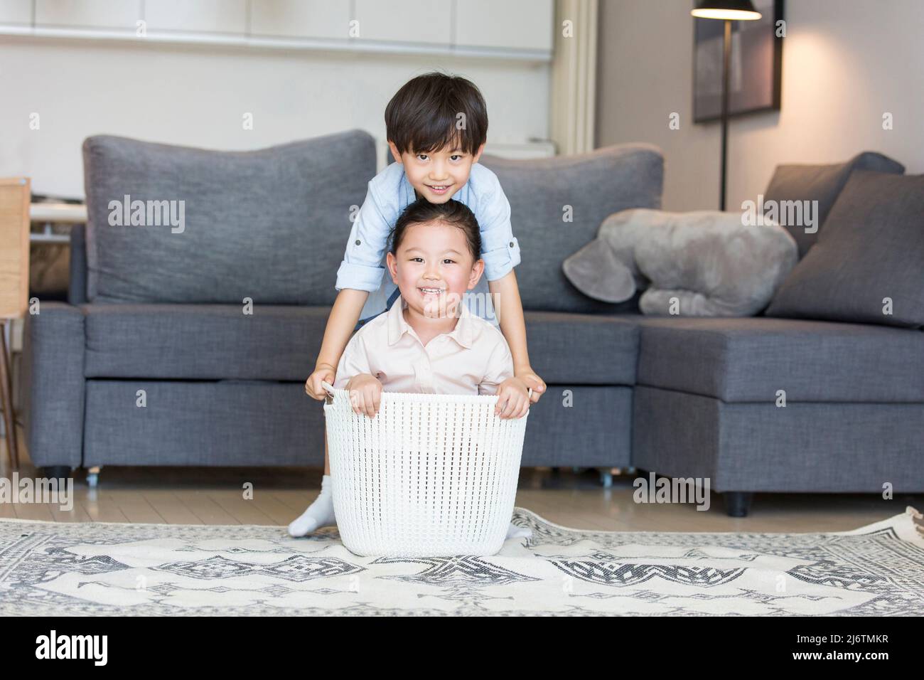 By the living room sofa, a little girl was ready to sit in the clothes basket and the little boy tried to move her up Stock Photo