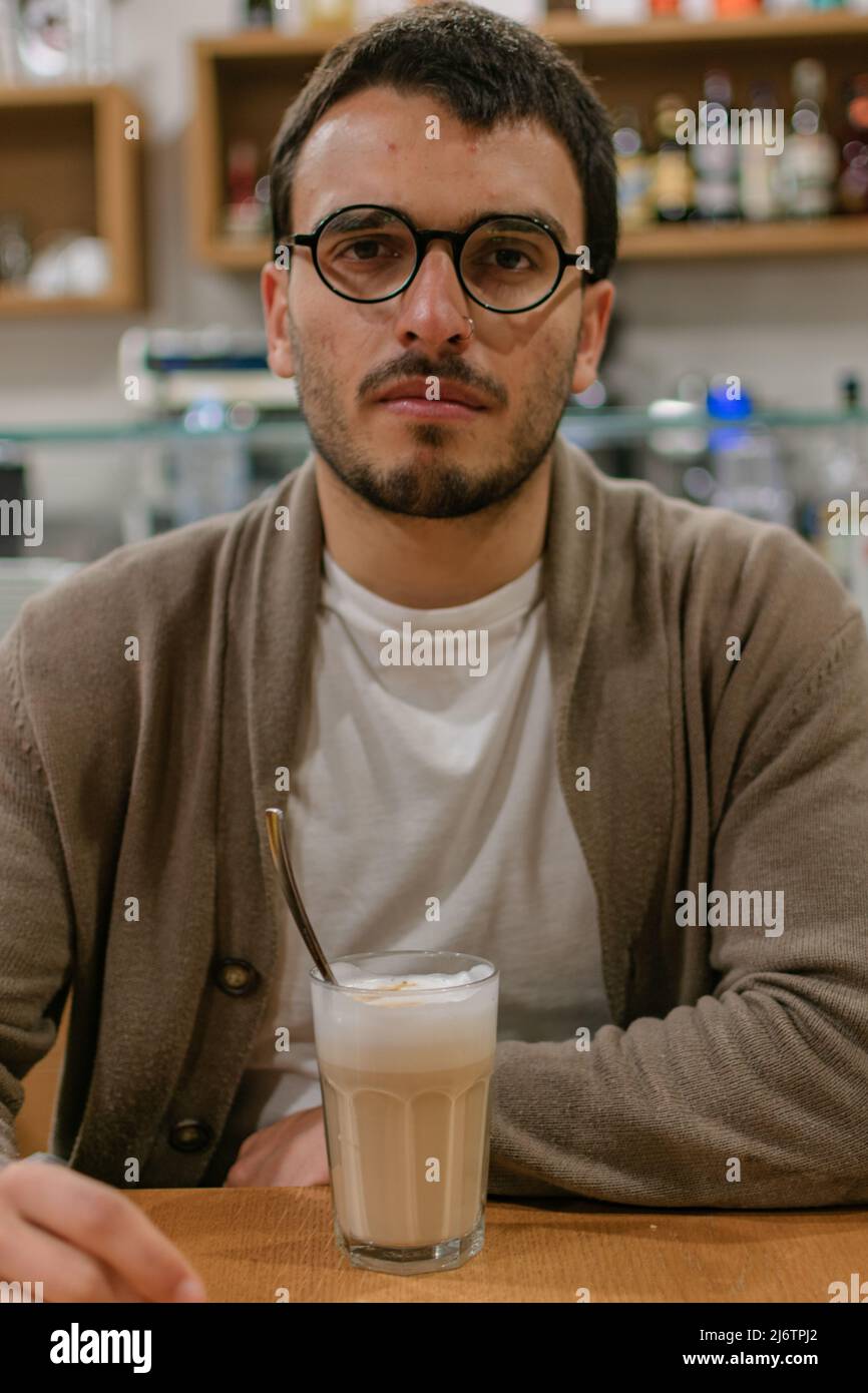Young tan bearded serious man with a Latte Macchiato Milk Cofee on a Table, grabbing the beverage wearing shirt short sleeve arms Bergamo Shop Italy Stock Photo