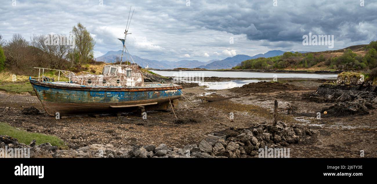 An abandoned boat pulled up onto the mud near Eilean Iarmain on Skye with the lighthouse at Eilean Sionnach visible in the background. Stock Photo