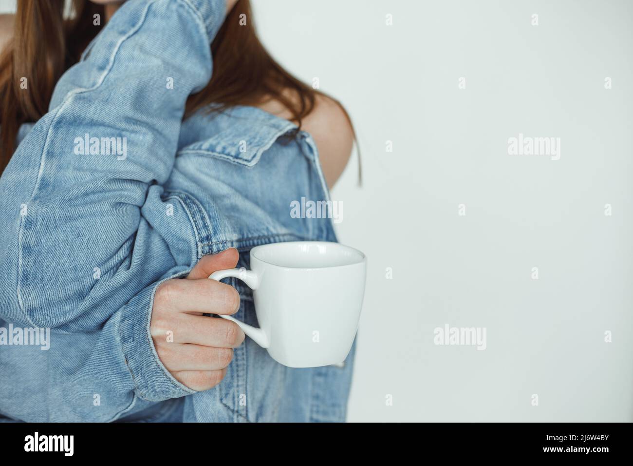 Cropped photo of woman with long dark hair wearing blue denim jacket backwards, holding white cup of coffee, posing. Stock Photo