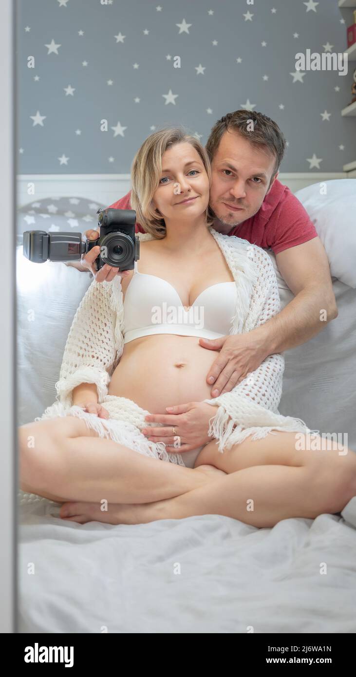 Pregnant couple taking selfie in mirror with digital camera. Husband embraces his wife. Family - man and woman. Pregnancy, maternity concept. Stock Photo