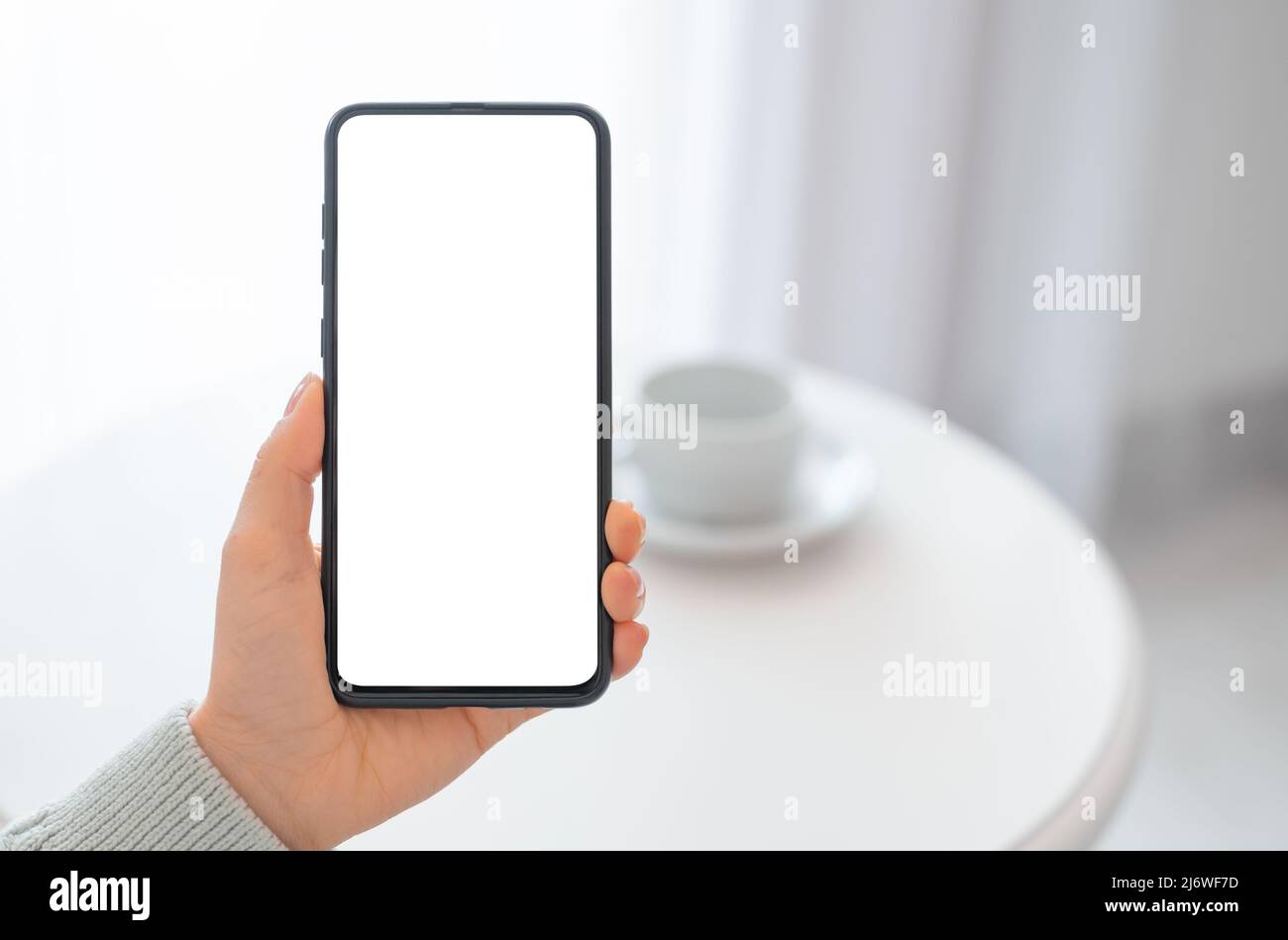 Mockup image woman holding black mobile phone with blank screen with coffee cup on table. woman's hand holding cellphone with blank desktop screen. To Stock Photo