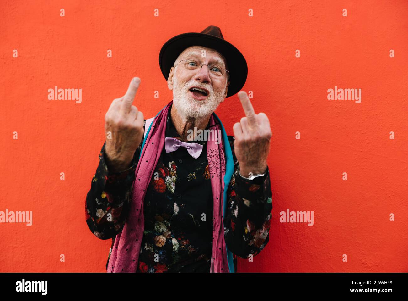 Happy eccentric man showing his middle fingers while standing against a red background. Mature man looking at the camera while making a swear gesture. Stock Photo