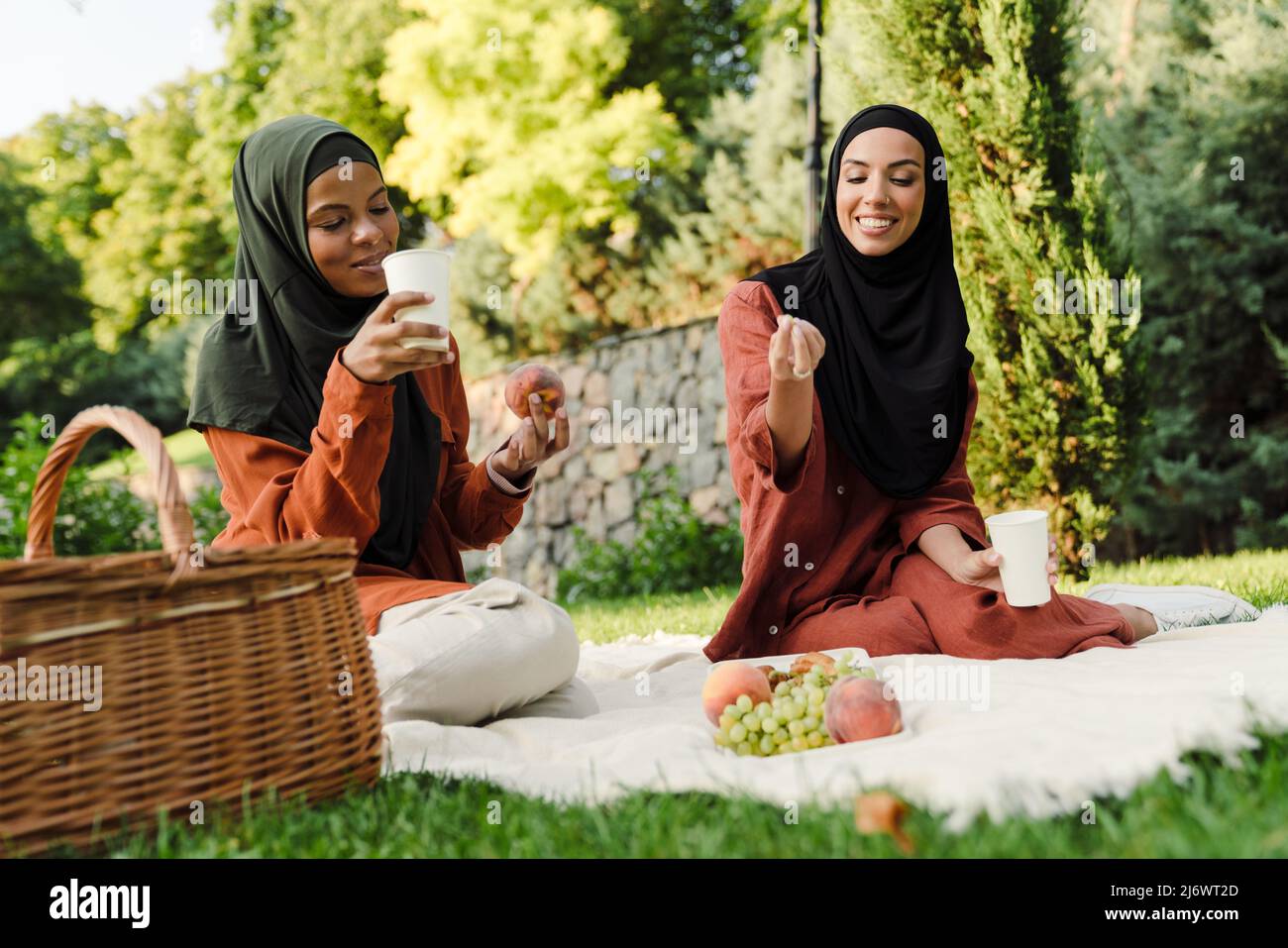 Multiracial muslim women smiling and eating fruits during picnic at green park Stock Photo