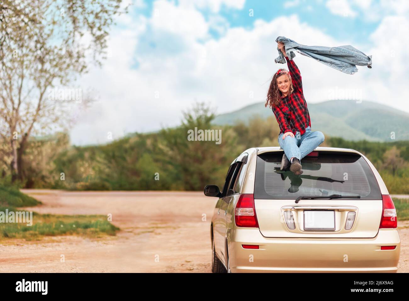Happy teen girl is sitting on the roof of a car, joyfully raising her ...