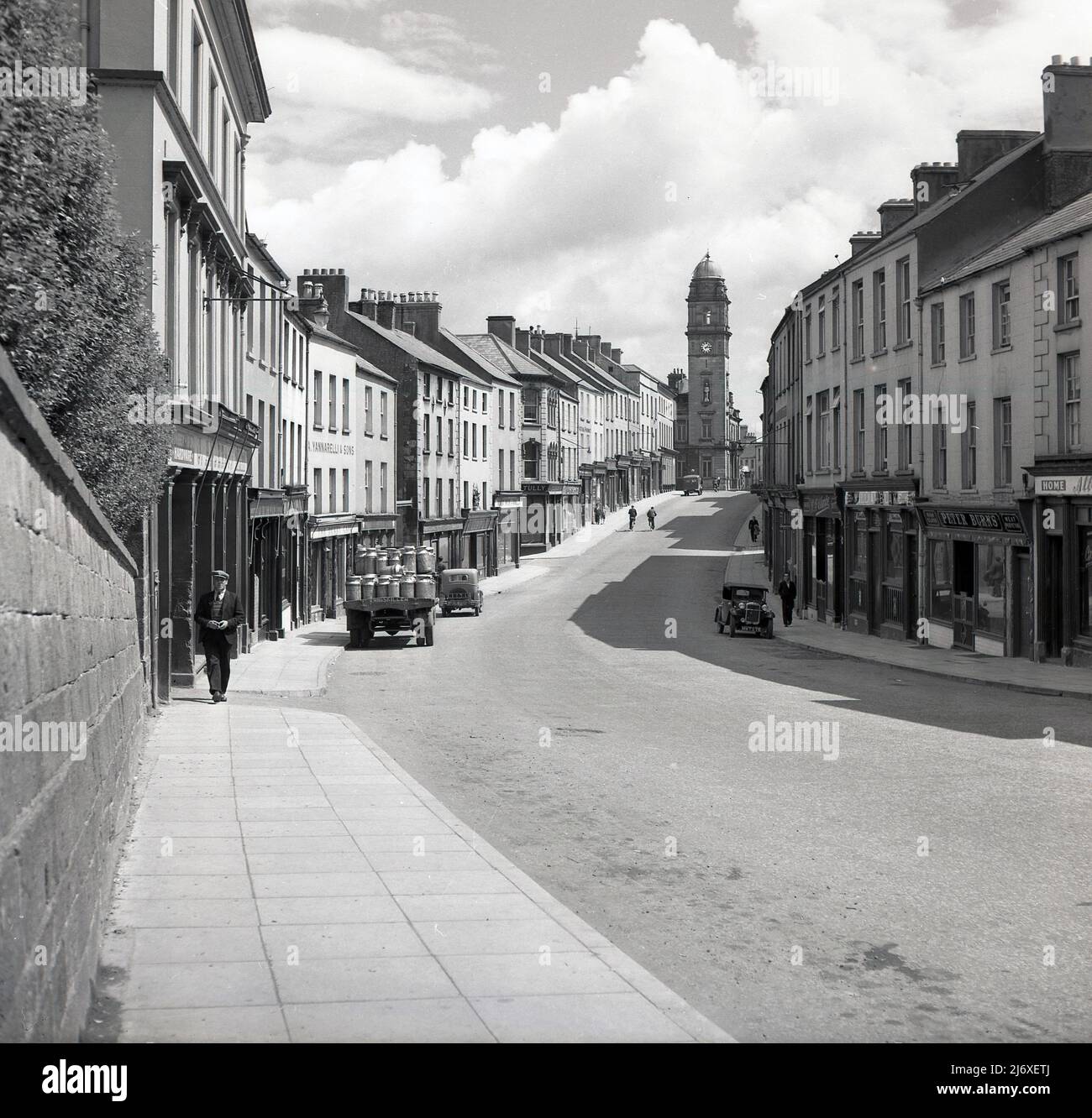 1950s, historical, Enniskillen, Northern Ireland, UK, showing a near deserted high street of with only a couple of locals walking along the pavement.  An open back truck with milk urns is parked up. Dating back to 1901, the clock tower of the town hall can be seen at the far end of the street. Names on stores include, a bar, William Blake; and a butcher and meat purveyor Peter Burns. The number plate on one of the cars parked in the deserted street, IL 2202, while on the other side of the street, another car, of an even earlier era is parked and this has the numberplate HMT 275. Stock Photo