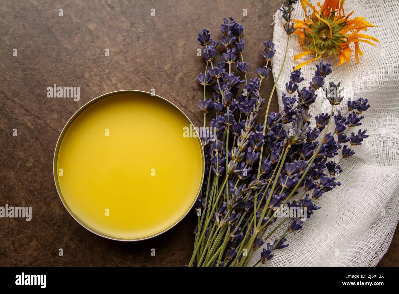 Closeup image taken from above of a tin of homemade DIY calendula and lavender salve next to the dried herbs and cheesecloth on brown tile background. Stock Photo