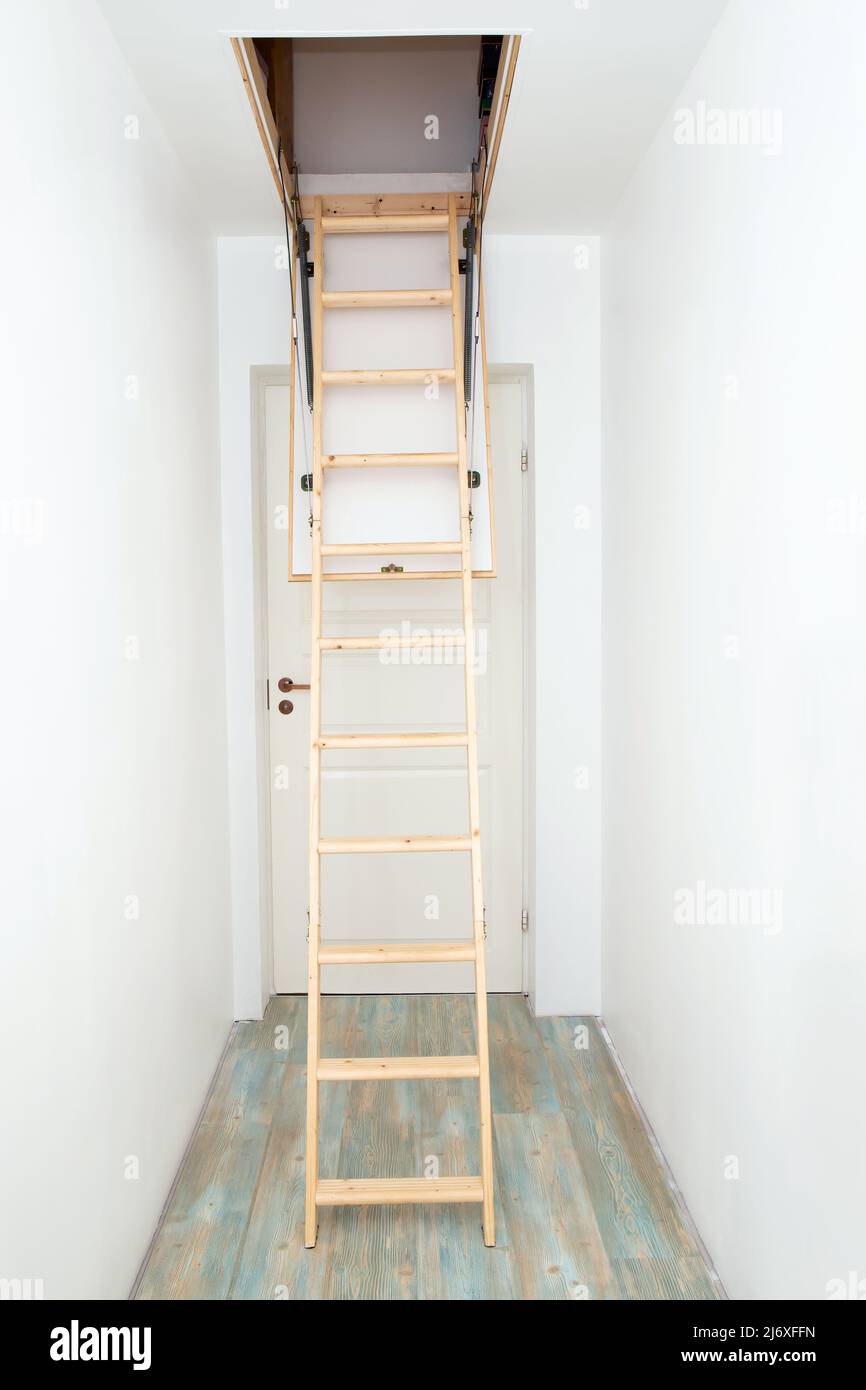Wooden foldable pull up attic stairs ladder at empty white home corridor. Narrow hallway with white walls and blue laminate flooring. Stock Photo