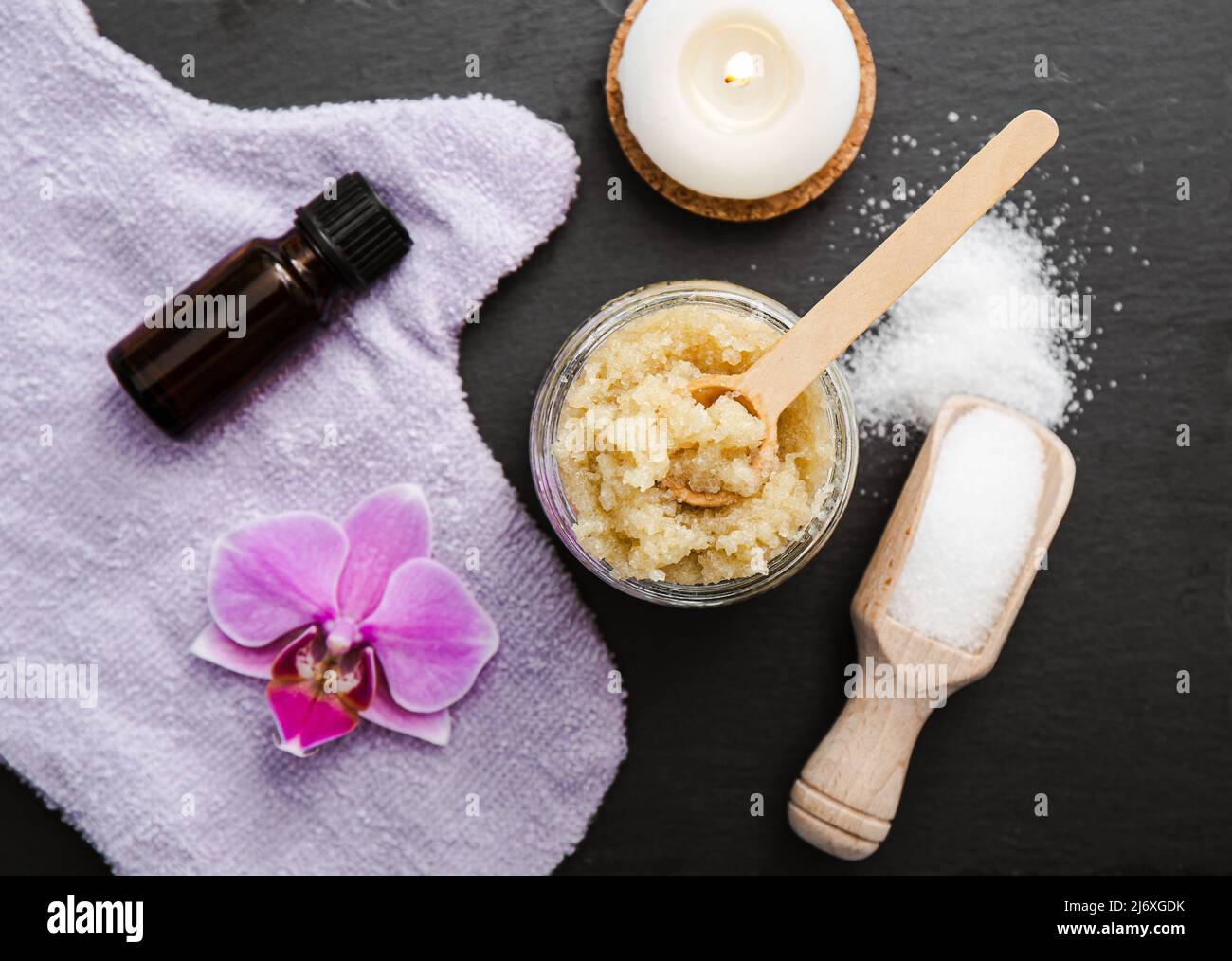 Homemade sugar body scrub in glass jar, decorated with orchid blossom, body scrub glove wooden spoon with sugar powder on black stone cutting board. Stock Photo