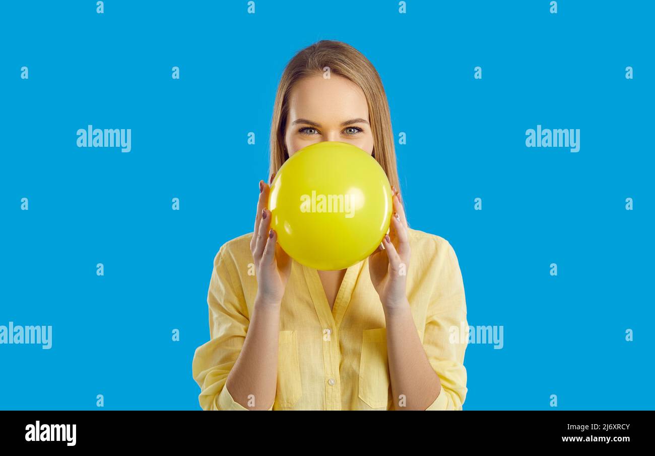 Studio portrait of happy young woman inflating yellow balloon and looking at camera Stock Photo