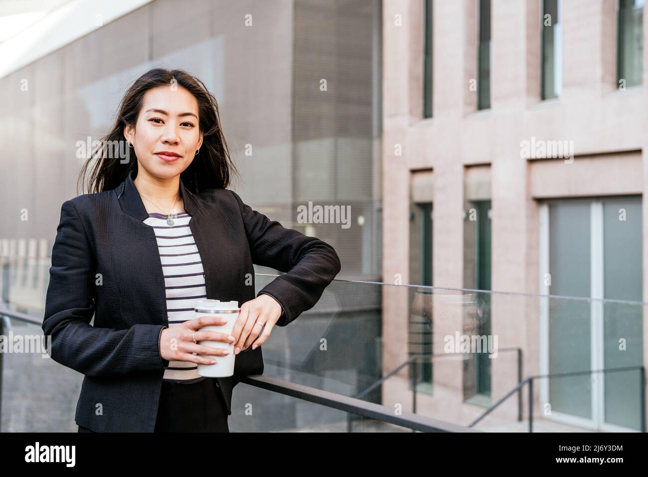 Young ethnic elegant female executive worker in black suit standing with zero waste tumbler bottle for coffee break in office district Stock Photo