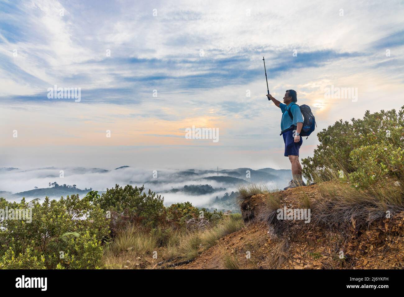 Hiker Asian man standing on peak of mountain with cloud scape raises hand to signify success Stock Photo