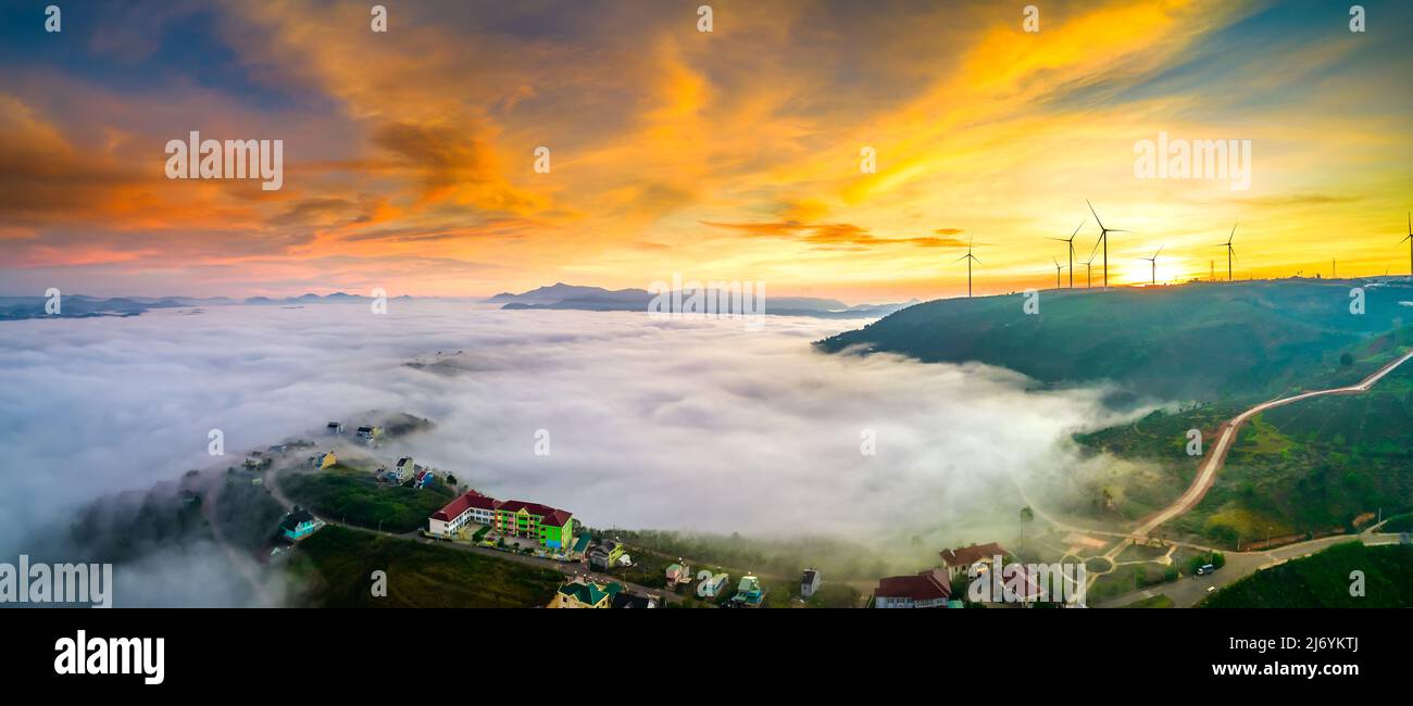 Aerial view top hill at dawn with fog covering small village in valley, beautiful wind power poles rising high to welcome a peaceful new day Stock Photo