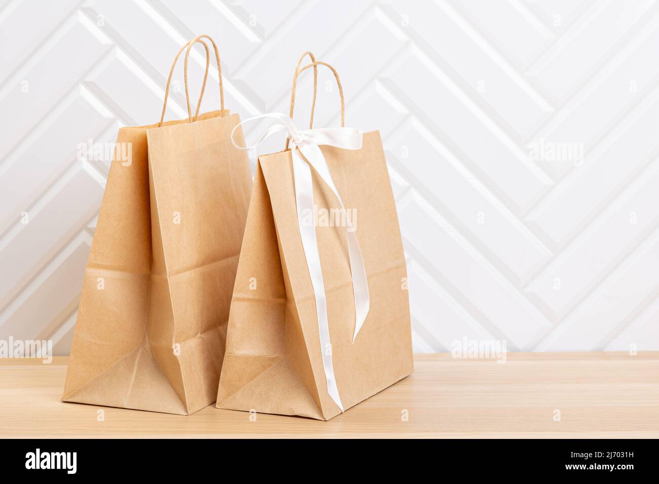 Blank  paper carrier bag with handles for shopping, facing front on right side of a wood veneer table with white wall background providing copy space Stock Photo