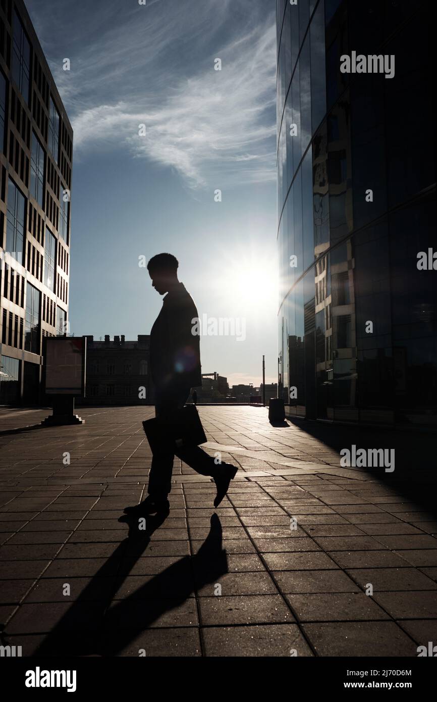 Working from dawn to dusk. Silhouette of male office worker with case going home after hard day at work, modern business centers seen behind him Stock Photo