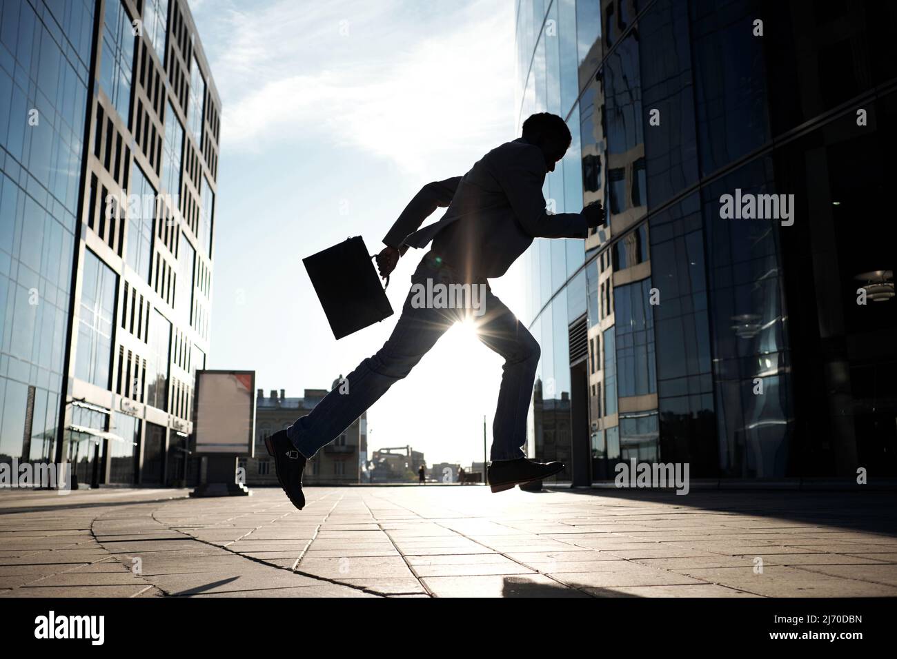 Silhouette of African American white-collar worker with case running towards modern office building against bright sun Stock Photo