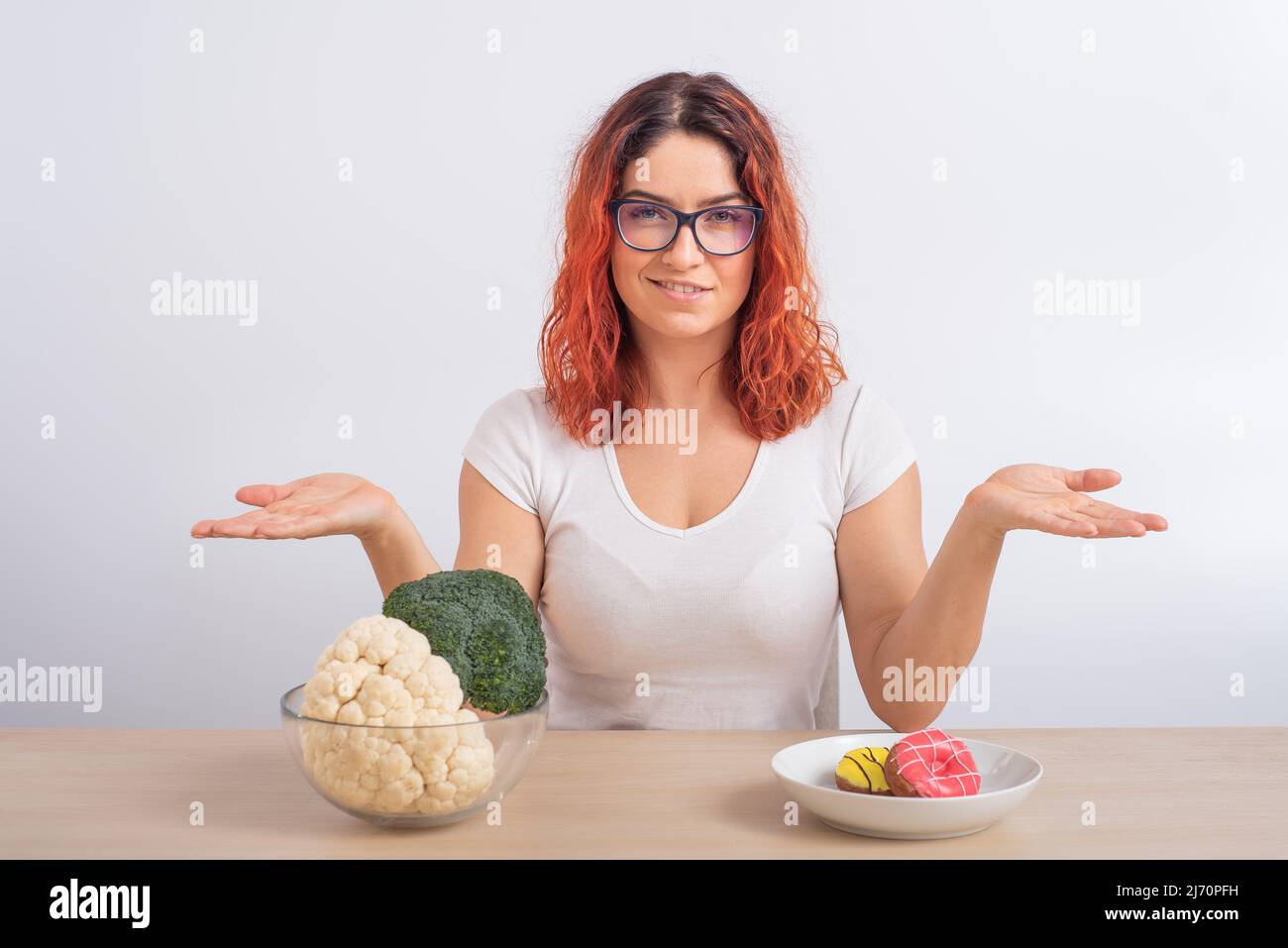 Caucasian woman prefers healthy food. Redhead girl chooses between broccoli and donuts on white background. Stock Photo