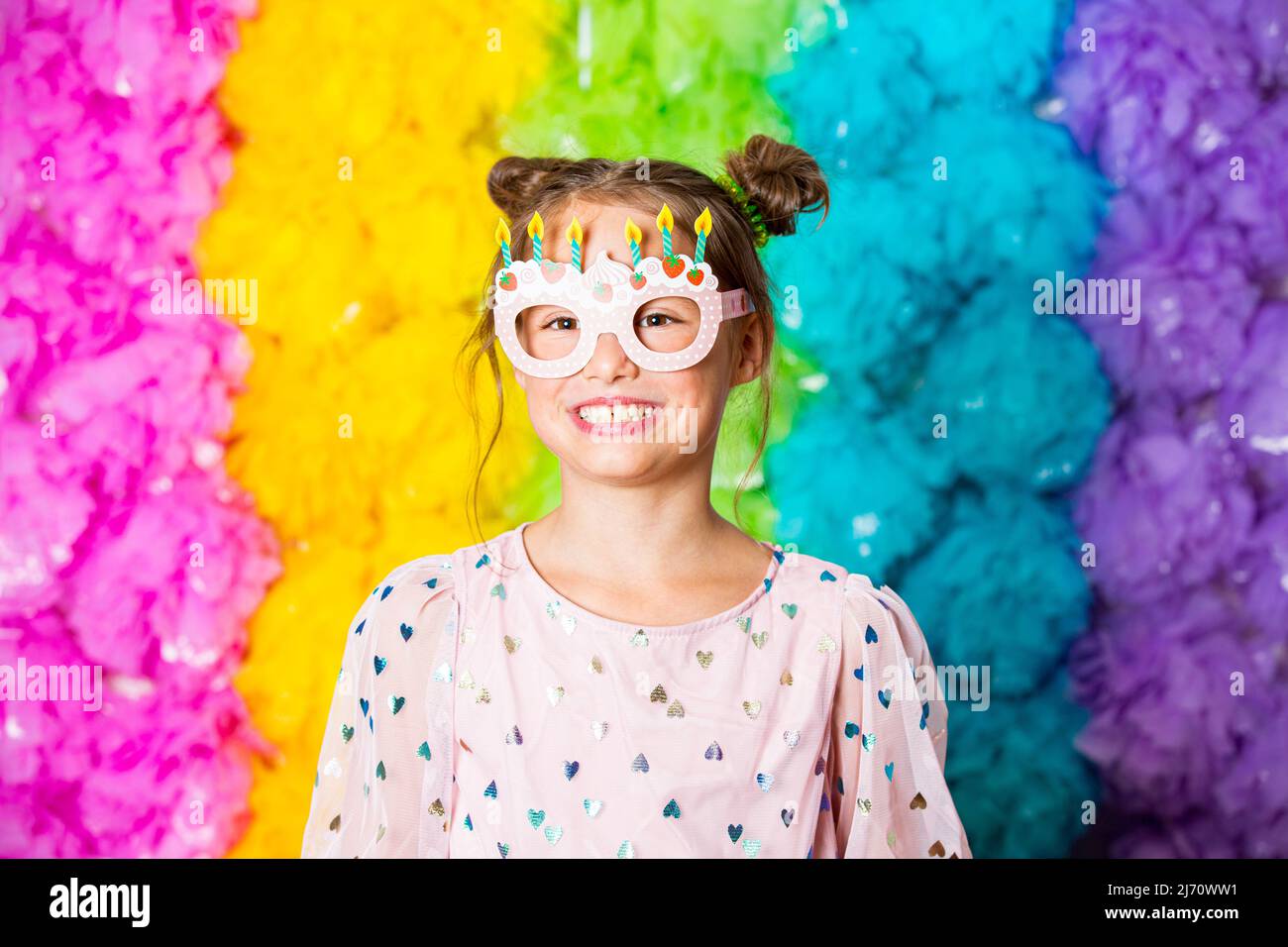 Cute girl celebrating birthday, wearing party paper glasses, laughing and having fun. Bright rainbow coloured background. Happy event. Stock Photo