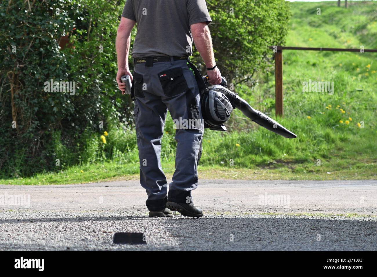 A man using a leaf blower on a driveway blower out onto a rural lane Stock Photo