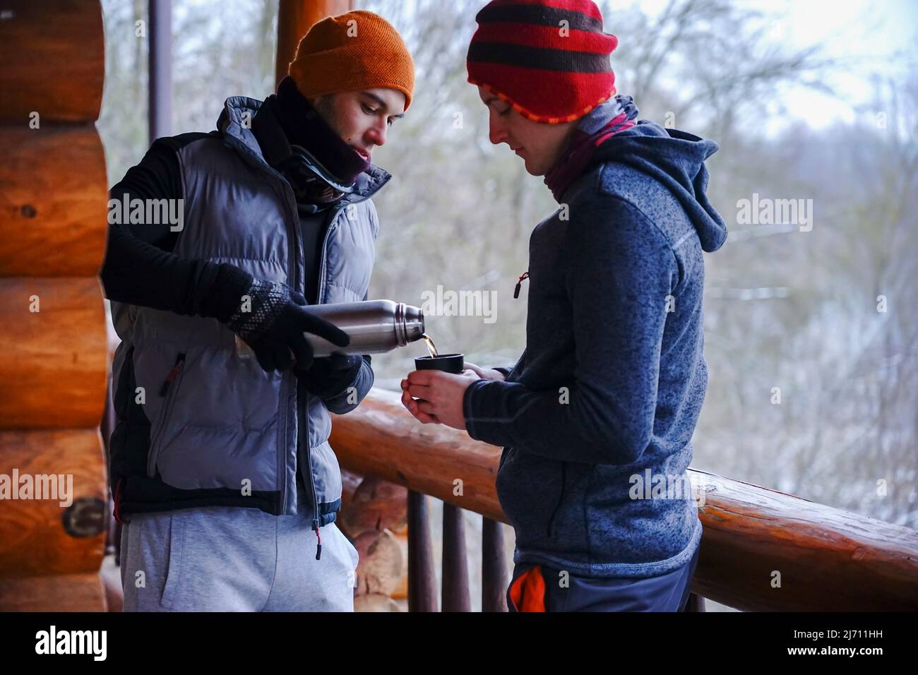 Two guys chat after skiing. They stand on the balcony of a wooden house in winter clothes, drink tea and relax. Porter of two guys in a ski resort in Stock Photo