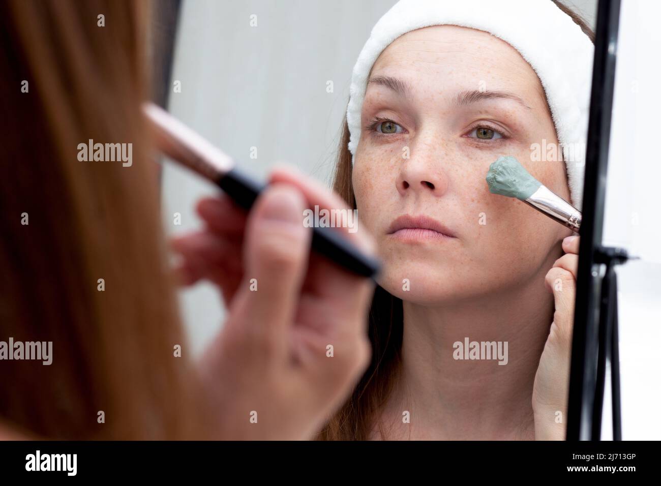 Caucasian middle-aged woman with white band on head putting blue clay on face using brush looking at herself in mirror Stock Photo
