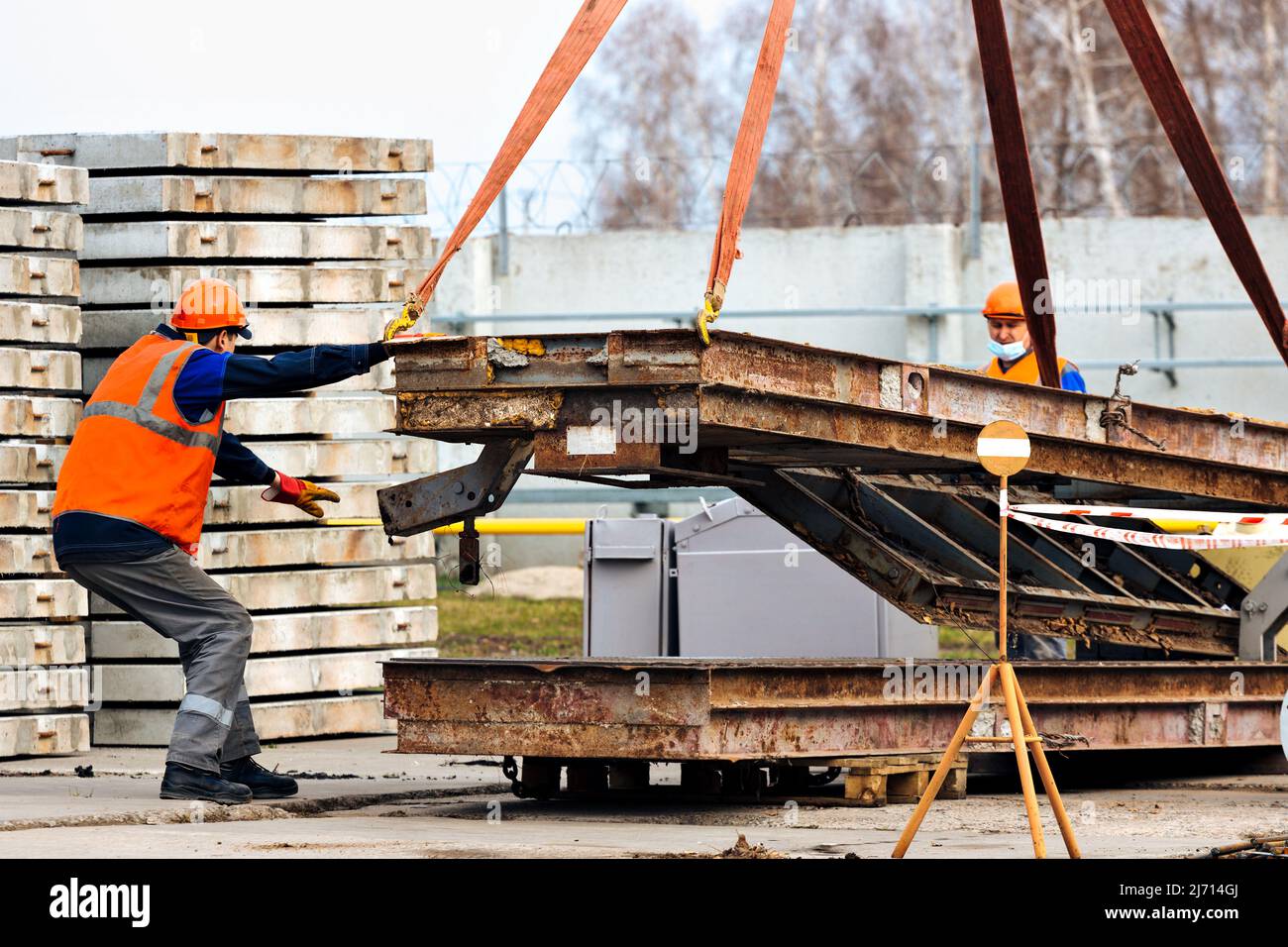 Slinger in helmet and vest controls unloading of metal structures on construction site. White handyman unloads load. Stock Photo