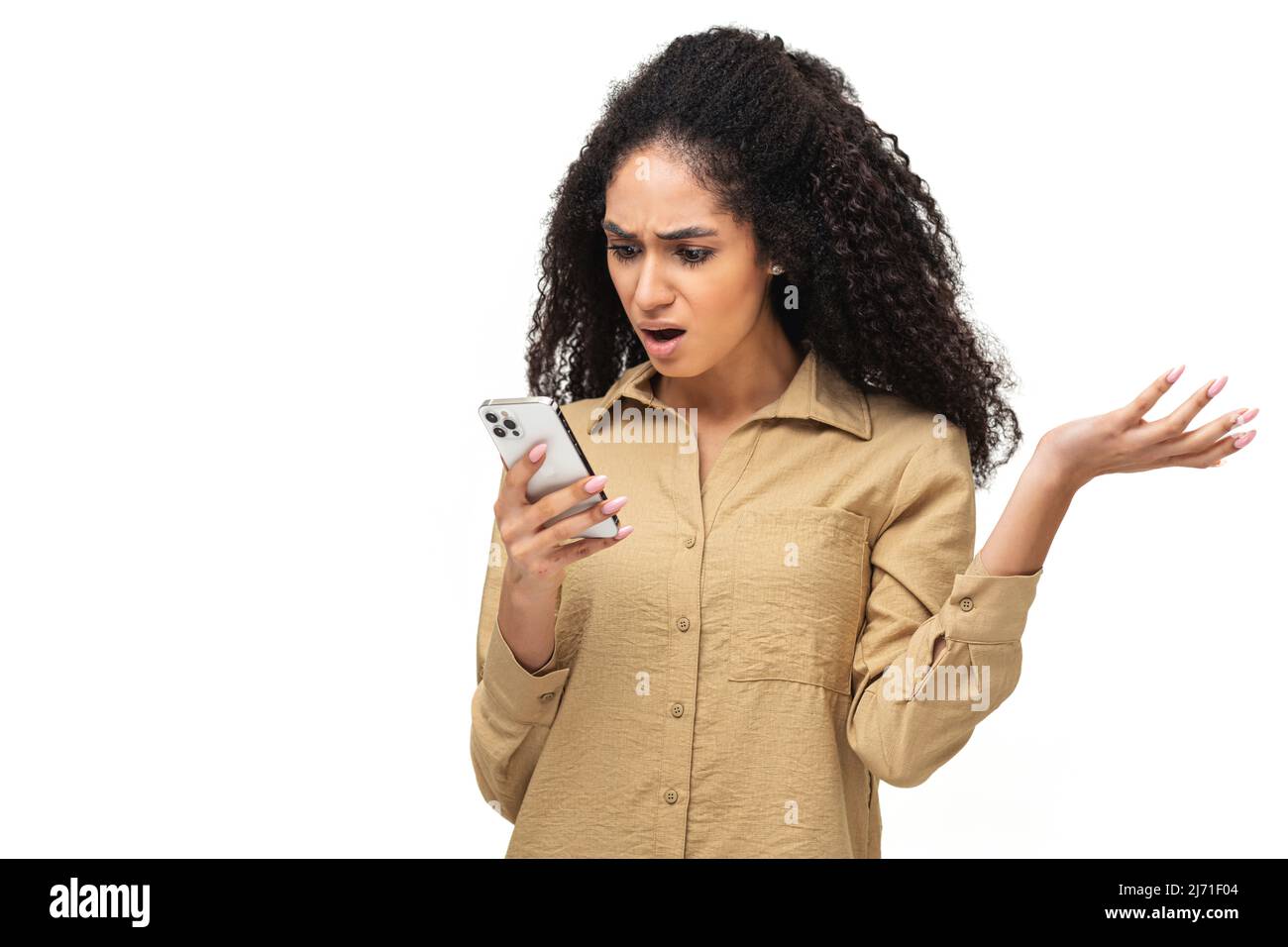 Disappointed young African-American woman holding smartphone and watching at the phone screen, lady received bad notification or messages, feels upset, shrugging, isolated Stock Photo