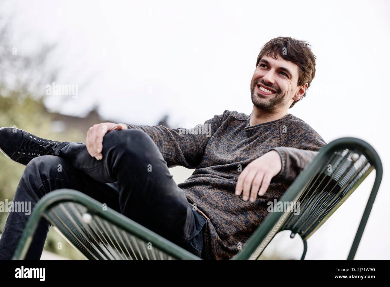 Junger Mann lachend draussen auf einer Parkbank sitzend, Portrait, Koeln, Nordrhein-Westfalen, Deutschland Stock Photo