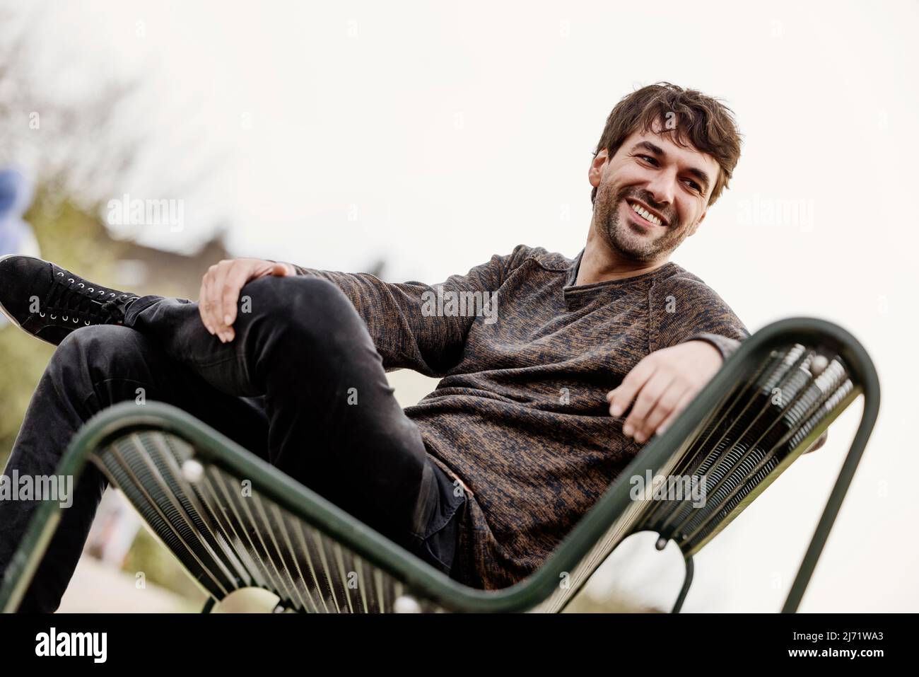 Junger Mann lachend draussen auf einer Parkbank sitzend, Portrait, Koeln, Nordrhein-Westfalen, Deutschland Stock Photo