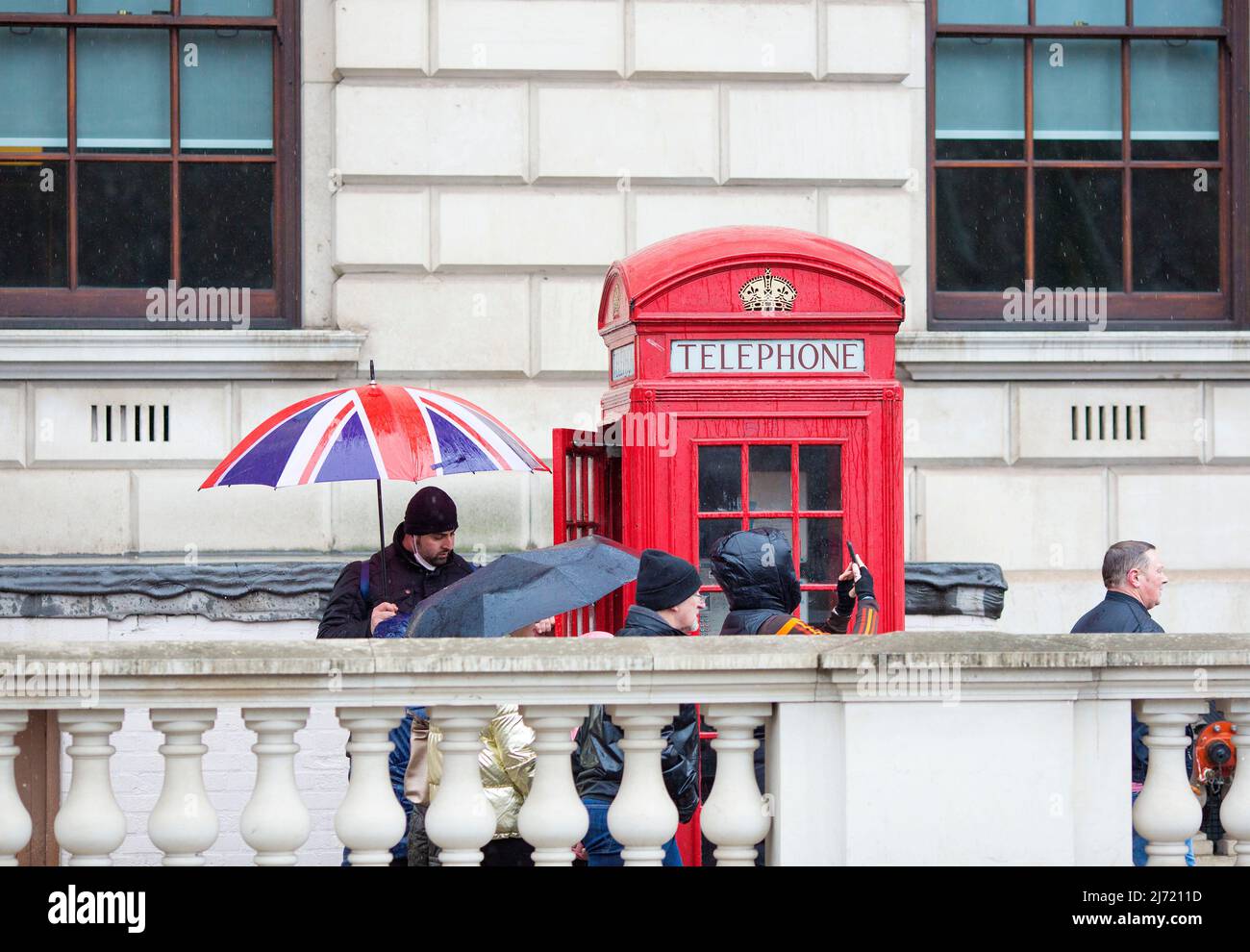 Pedestrians walk past a person holding a Union flag design umbrella next to a traditional red telephone box in Westminster, central London. Stock Photo